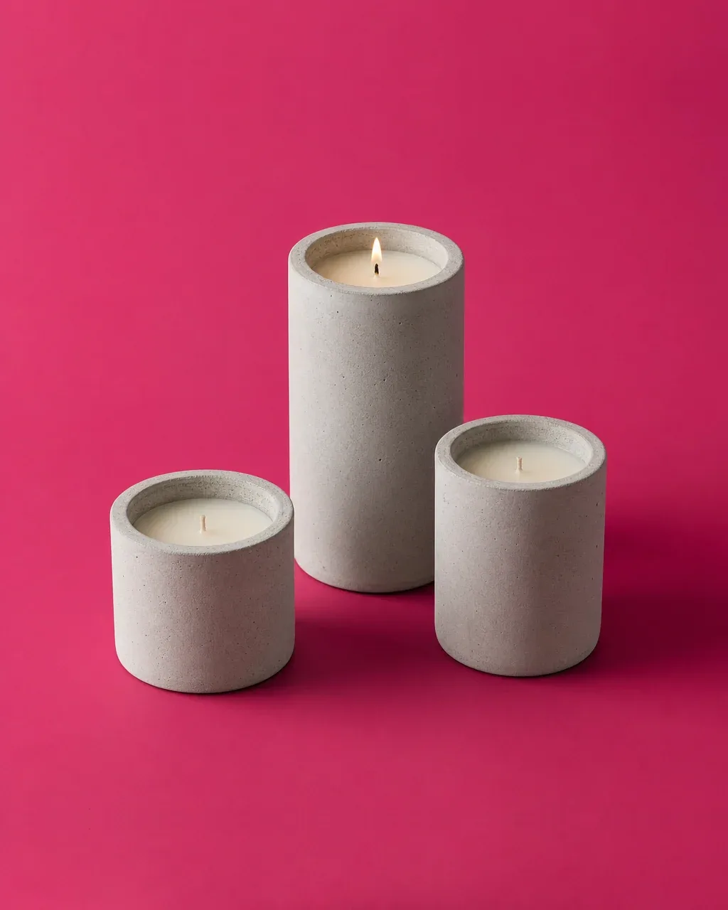 Recipientes cilíndricos de concreto para vela de distintas alturas, uno encendido, sobre fondo magenta.